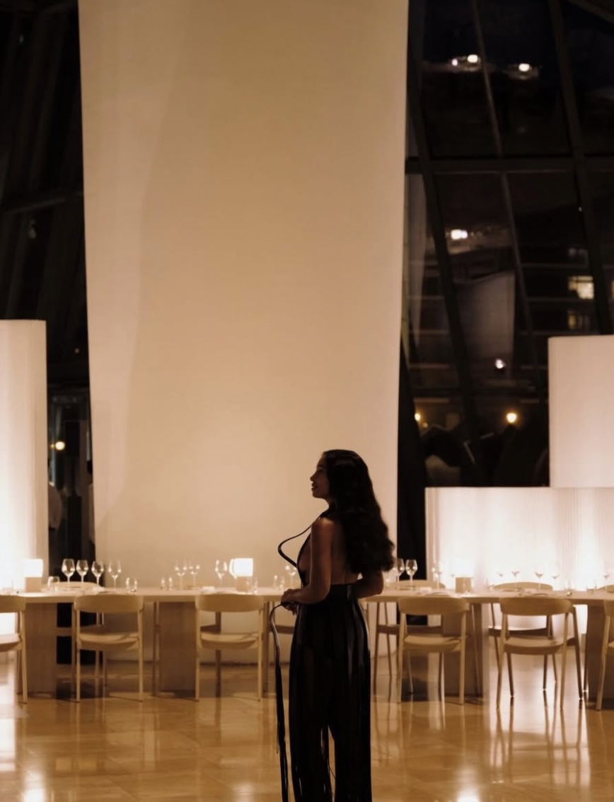 Candlelit dining hall — a woman in black, warm light, long set tables